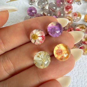 Puede incluir: Primer plano de varias esferas pequeñas de resina transparente, cada una encapsulando una flor seca. Las flores son de varios colores, incluyendo blanco, rojo, amarillo y morado. Algunas esferas tienen bucles plateados.