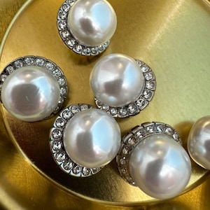 May include: A close-up of six white pearl buttons with a silver rhinestone border. The buttons are on a gold surface.