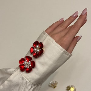 May include: Two red flower-shaped cufflink sets with pearl accents are attached to a white shirt cuff. The cufflinks have a glossy finish. Two additional flower-shaped accessories are visible in the background.