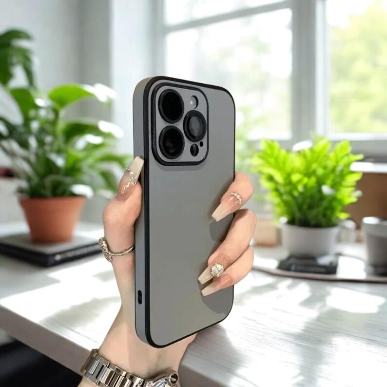K&ouml;nnte beinhalten: Eine graue Smartphone-H&uuml;lle mit schwarzem Rand. Die H&uuml;lle hat eine matte Oberfl&auml;che und einen Kameraausschnitt. Die Handyh&uuml;lle wird von einer Person mit langen N&auml;geln und einer silbernen Uhr gehalten.