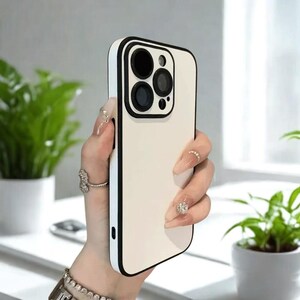 K&ouml;nnte beinhalten: Eine cremefarbene Handyh&uuml;lle mit schwarzem Rand. Die H&uuml;lle hat einen Kameraausschnitt mit schwarzen Kreisen. Die Handyh&uuml;lle wird in einer Hand mit langen, gepflegten N&auml;geln und Ringen gehalten.