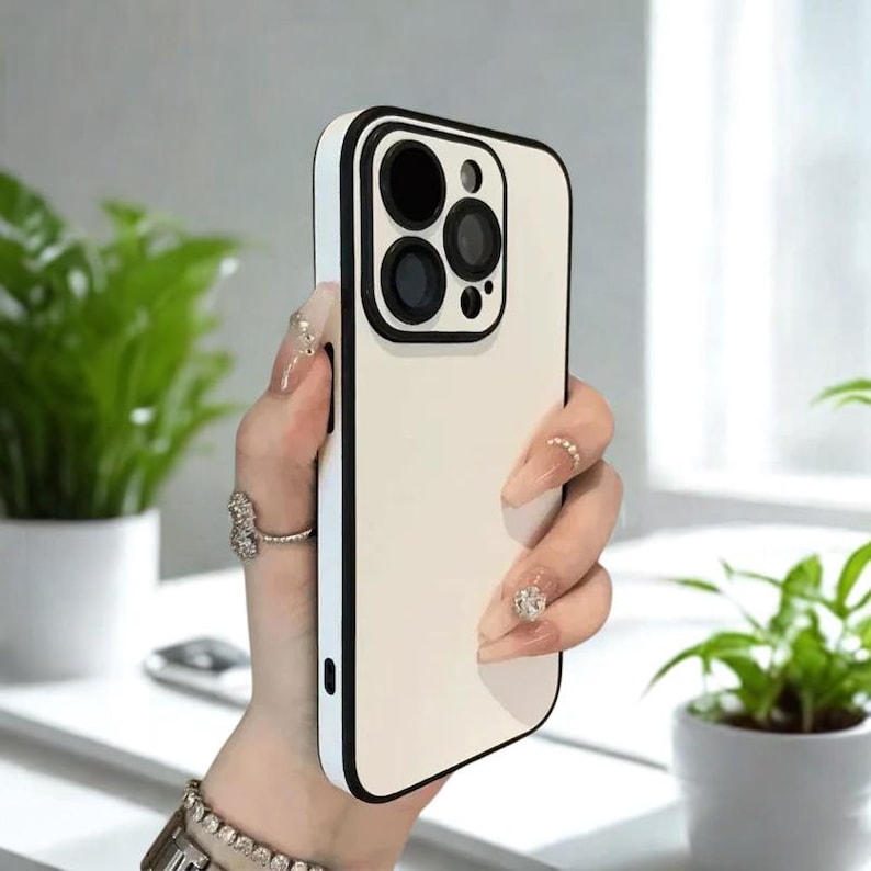 K&ouml;nnte beinhalten: Eine cremefarbene Handyh&uuml;lle mit schwarzem Rand. Die H&uuml;lle hat einen Kameraausschnitt mit schwarzen Kreisen. Die Handyh&uuml;lle wird in einer Hand mit langen, gepflegten N&auml;geln und Ringen gehalten.
