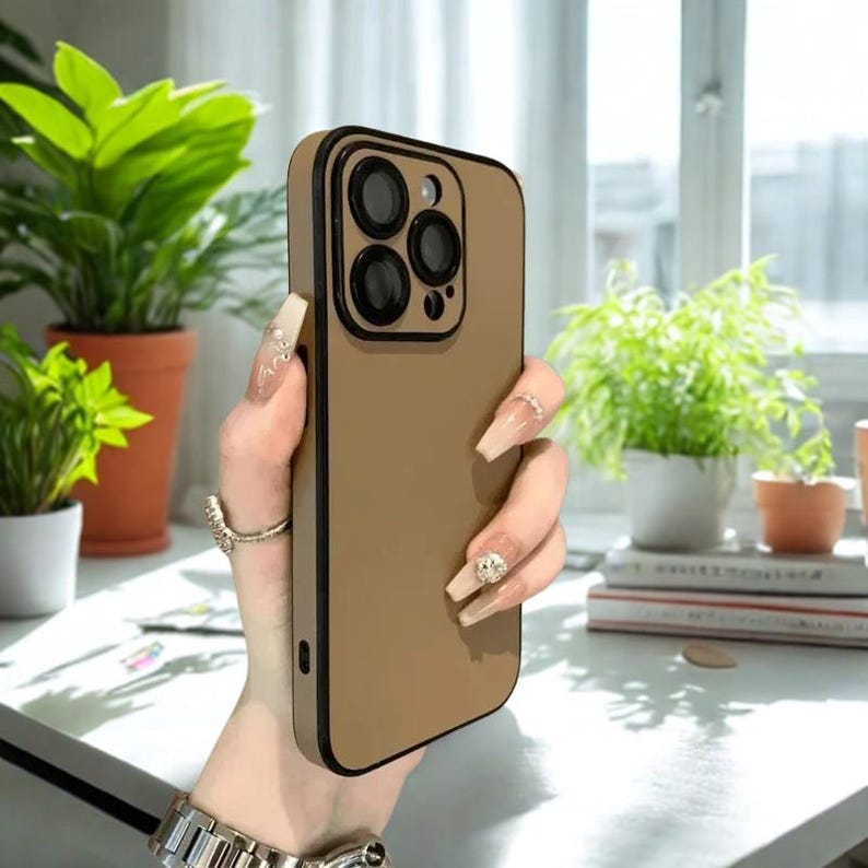 K&ouml;nnte beinhalten: Eine hellbraune Handyh&uuml;lle mit schwarzem Rand und Kamerarahmen. Die H&uuml;lle hat eine glatte Oberfl&auml;che und sch&uuml;tzt das Telefon. Die H&uuml;lle wird in einer hellen Umgebung mit Pflanzen im Hintergrund gezeigt.