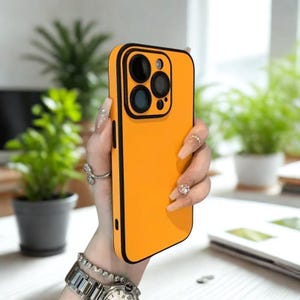 K&ouml;nnte beinhalten: Eine leuchtend orangefarbene Handyh&uuml;lle mit schwarzem Rand. Die H&uuml;lle hat eine Kamera&ouml;ffnung mit drei Linsen. Die Handyh&uuml;lle wird vor einem verschwommenen Hintergrund mit Pflanzen gehalten.