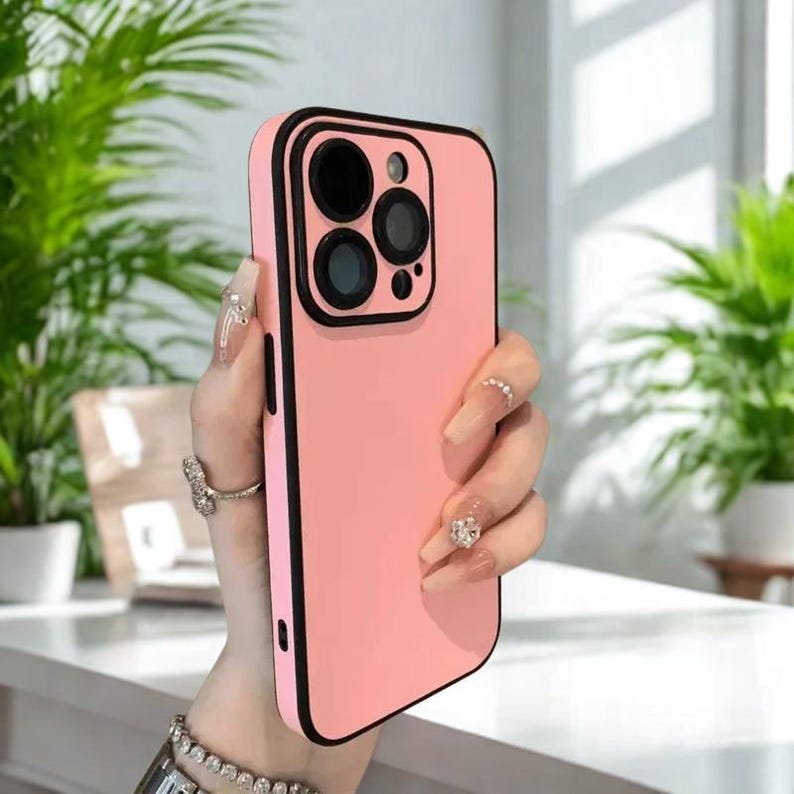 K&ouml;nnte beinhalten: Eine rosa Handyh&uuml;lle mit schwarzem Rand und Kameradetails. Die H&uuml;lle wird in einer Hand gehalten, wodurch ihr schlankes Design und ihre Schutzeigenschaften hervorgehoben werden. Die H&uuml;lle ist f&uuml;r ein Smartphone konzipiert.