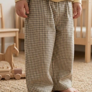 Puede incluir: Pantalones largos con un patrón de cuadros en beige y negro. Los pantalones tienen un corte recto con una cintura elástica fruncida y se combinan con un suéter de punto de manga larga en color amarillo claro metido por dentro.