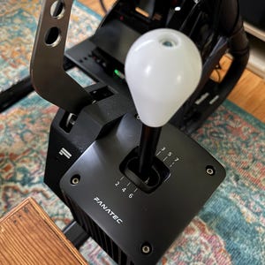 Könnte beinhalten: Ein schwarzer Fanatec Rennsimulator-Schalthebel mit einem weißen Schaltknauf. Der Schalthebel hat Ganganzeigen und ist auf einem schwarzen Metallrahmen montiert. Das Bild zeigt einen Holztisch und einen bunten Teppich im Hintergrund.