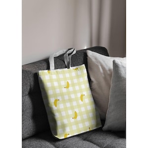 May include: A light green and white checkered tote bag with yellow banana illustrations. The bag has gray handles and is resting on a gray sofa next to striped and solid gray pillows. The bag is made of fabric.