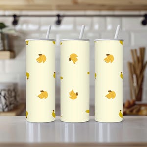 May include: Three cylindrical tumblers with white straws. The tumblers have a light yellow background and are decorated with a pattern of yellow bananas. The tumblers are made of stainless steel and are designed to keep drinks cold.