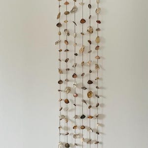 Op de afbeelding: Een decoratieve wandhanger gemaakt van verschillende schelpen. De schelpen zijn geregen aan dunne koorden en opgehangen aan een houten balk en een touw. De schelpen zijn in tinten beige, bruin en wit.