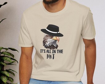 Puede incluir: Una camiseta beige con una ilustración en blanco y negro de un águila con un fedora. El texto "It's all in the HAT" está impreso debajo del águila.