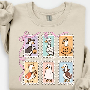 May include: Beige sweatshirt featuring six Halloween-themed goose illustrations in postage stamp frames. Each goose is dressed in a costume, including a witch, mummy, and ghost. A pink ribbon and decorative stars add to the festive design.