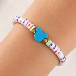 May include: A stretch bracelet featuring small, flat, multicolored disc beads in white, orange, purple, and green. The bracelet is accented with two small gold-toned metallic spheres flanking a central bright blue charm shaped like a mouse head.