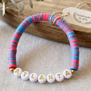 May include: A handmade bracelet featuring alternating pink and blue disc beads. The word "ATLANTA" is spelled out in white letter beads with gold lettering. A tag with the text "Handmade in ATL" is attached.