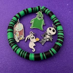 Puede incluir: Una pulsera de cuentas verdes y negras con cuatro colgantes con personajes de Pesadilla antes de Navidad. Los colgantes incluyen a Sally, Oogie Boogie, Zero y Jack Skellington. La pulsera está sobre una superficie morada.