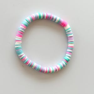 Könnte beinhalten: Ein farbenfrohes Armband aus flachen, runden Perlen. Das Armband weist eine Mischung aus Pastellfarben auf, darunter Rosa, Blau, Grün, Weiß und Lila. Die Perlen sind zu einer kreisförmigen Form aufgefädelt.