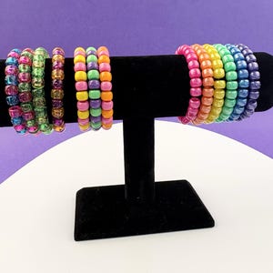 May include: A selection of beaded bracelets in various colours, displayed on a black velvet stand. The bracelets showcase a spectrum of hues, including pink, orange, yellow, green, and blue. The small, round beads create a lively appearance. The stand is on a white surface with a purple backdrop.