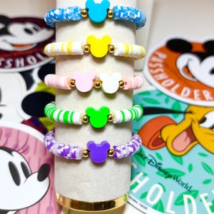 Könnte beinhalten: Fünf bunte Armbänder mit Mickey Mouse Kopf-Anhängern. Die Armbänder sind blau, gelb, rosa, grün und lila, mit goldfarbenen Perlen. Die Armbänder werden auf einem weißen Zylinder präsentiert.