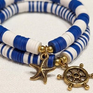 May include: Two nautical-themed bracelets. Each bracelet is made of blue and white disc-shaped beads. One bracelet has a gold starfish charm, and the other has a gold ship's wheel charm. The bracelets are on a white surface.