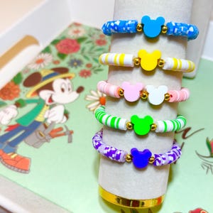 Könnte beinhalten: Eine Sammlung farbenfroher Perlenarmbänder mit Mickey-Mouse-Silhouetten. Die Armbänder sind in verschiedenen Farben gehalten, darunter Blau, Gelb, Rosa, Grün und Lila, mit goldfarbenen Akzenten. Die Armbänder werden auf einem weißen zylindrischen Ständer präsentiert.