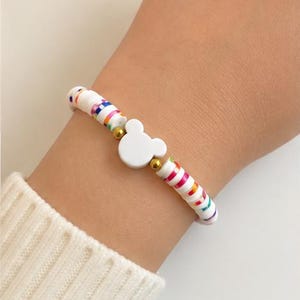 May include: A stretch bracelet featuring flat, circular white clay beads interspersed with vibrant, multi-colored striped beads. The design is centered with a solid white, rounded mouse-ear silhouette charm, flanked by two small, polished gold-tone metallic spacer beads.