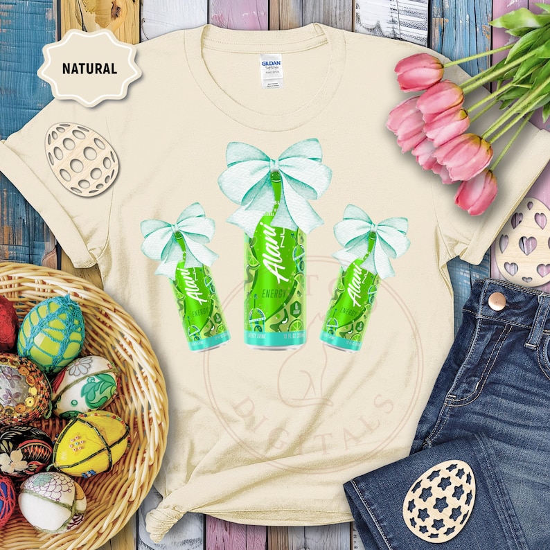 May include: A cream-colored t-shirt featuring a watercolor design of three green energy drink cans with light blue bows. The shirt is styled with a basket of decorated Easter eggs, pink tulips, and denim jeans.