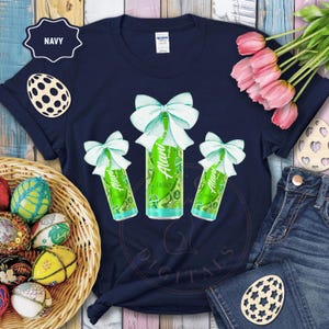 May include: Navy blue t-shirt featuring three green energy drink cans with light blue bows. The cans have the text "Alani Energy" and lime graphics. The shirt is decorated with Easter-themed elements, including decorative eggs and a wooden egg.