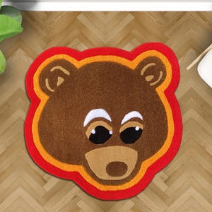 May include: A brown bear-shaped rug with a red, orange, and brown border. The rug features a bear face with white eyes, black pupils, and a tan snout. The rug is placed on a wooden floor.