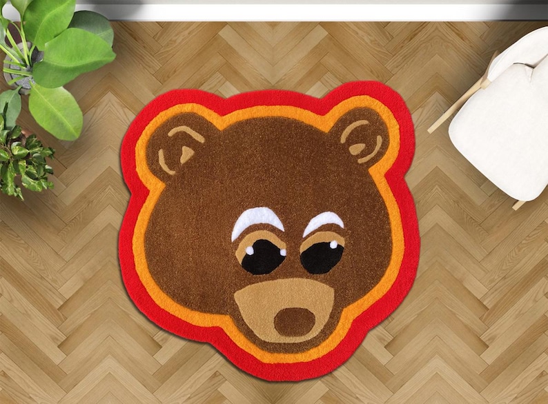 May include: A brown bear-shaped rug with a red, orange, and brown border. The rug features a bear face with white eyes, black pupils, and a tan snout. The rug is placed on a wooden floor.