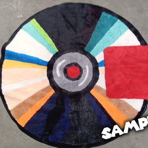 May include: A round rug designed to resemble a vinyl record. The rug features a black center with a red dot, surrounded by a gray ring. The outer section is divided into colorful segments, including blue, orange, and white. A red square is on the right side.