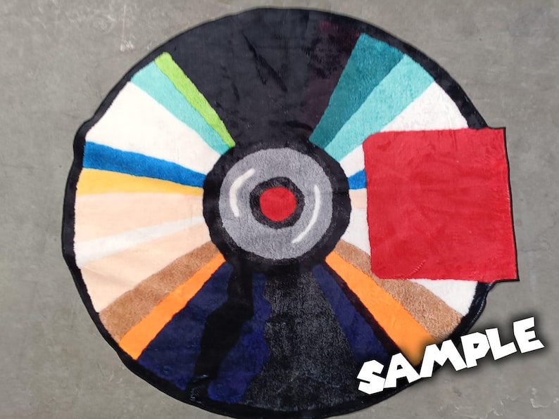 May include: A round rug designed to resemble a vinyl record. The rug features a black center with a red dot, surrounded by a gray ring. The outer section is divided into colorful segments, including blue, orange, and white. A red square is on the right side.