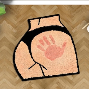 May include: A novelty bath mat shaped like a rear end, featuring a peach-colored design with a black waistband and outline. A pink handprint is centered on the mat. The mat is placed on a wooden floor.