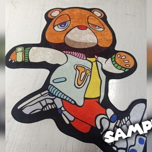 May include: A colorful rug featuring a cartoon bear with a sad expression. The bear is wearing a jacket with a "T" on it, and the design incorporates elements of a skeleton. The rug is approximately 48 inches long.