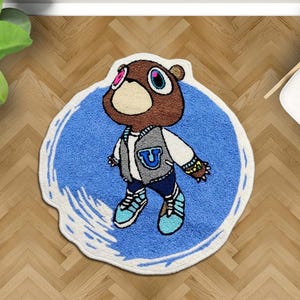 May include: A cartoon-style rug featuring a brown bear character wearing a gray and white varsity jacket with a blue "U" on the chest. The rug has a blue background with a white border and is placed on a wooden floor.
