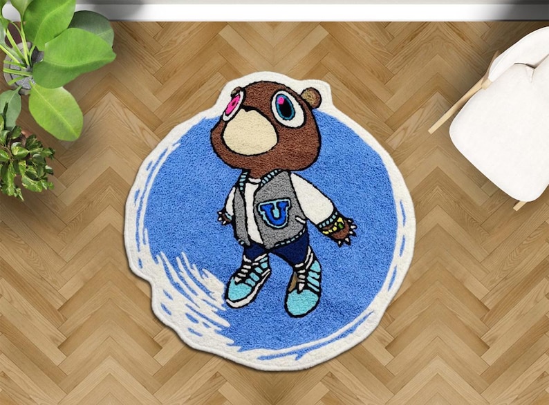 May include: A cartoon-style rug featuring a brown bear character wearing a gray and white varsity jacket with a blue "U" on the chest. The rug has a blue background with a white border and is placed on a wooden floor.