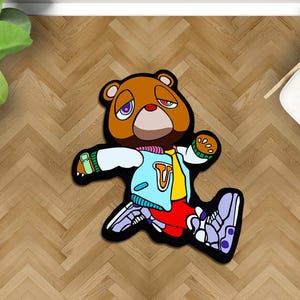 May include: A brown bear-shaped rug with a cartoon design. The bear wears a blue jacket, red pants, and purple sneakers. The rug has a black border and is placed on a wooden floor.