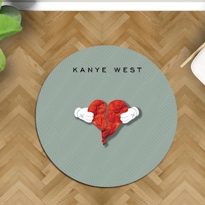 May include: A round, light teal rug with the text "KANYE WEST" in black. The rug features a red, broken heart graphic held by white cartoon hands with black outlines. The rug is on a wooden floor.