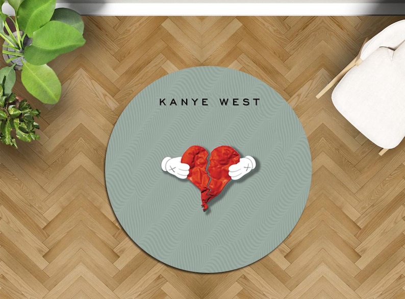 May include: A round, light teal rug with the text "KANYE WEST" in black. The rug features a red, broken heart graphic held by white cartoon hands with black outlines. The rug is on a wooden floor.