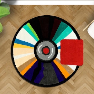 May include: A colorful, circular rug with a black border, resembling a vinyl record. The rug features a variety of colors, including red, white, blue, and green. A small, red square rug is placed next to the circular rug. The rug is on a wooden floor.