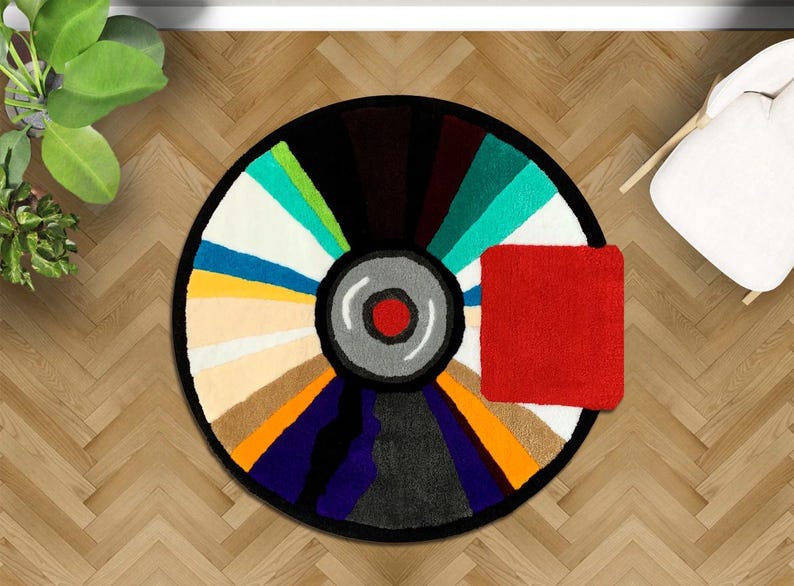 May include: A colorful, circular rug with a black border, resembling a vinyl record. The rug features a variety of colors, including red, white, blue, and green. A small, red square rug is placed next to the circular rug. The rug is on a wooden floor.