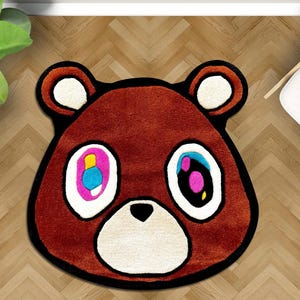 May include: Brown bear-shaped rug with a black outline and white accents. The bear's eyes are colorful, with pink, blue, and yellow details. The rug is on a wooden floor, and a white chair is visible in the background.