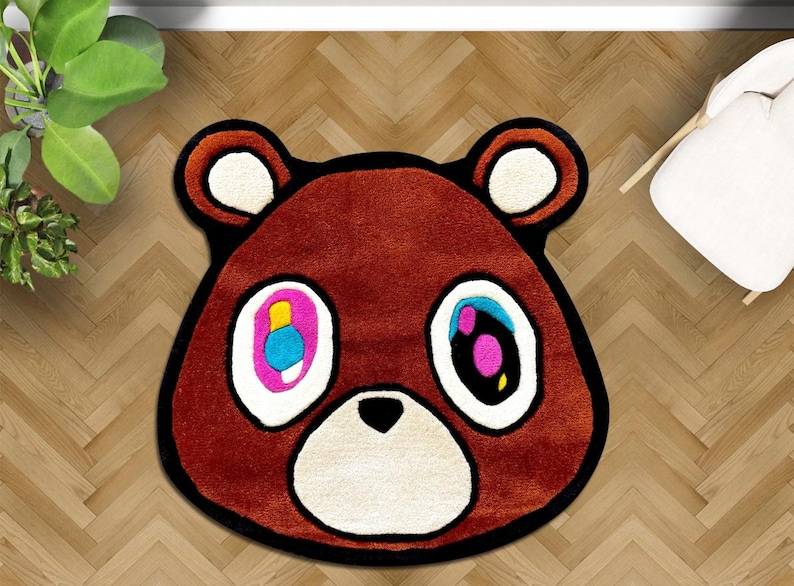 May include: Brown bear-shaped rug with a black outline and white accents. The bear's eyes are colorful, with pink, blue, and yellow details. The rug is on a wooden floor, and a white chair is visible in the background.