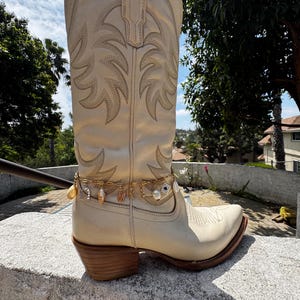 Può includere: Stivali da cowboy bianchi con una catena dorata e ciondoli intorno alla caviglia. I ciondoli includono conchiglie, una perla e una carta da gioco.