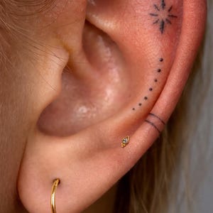 Puede incluir: Una oreja humana con un pequeño pendiente de aro dorado en el lóbulo y un diminuto pendiente de botón dorado en la concha. El cartílago superior muestra un delicado tatuaje de tinta negra con un símbolo de estrella, puntos y dos líneas.