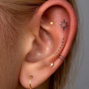 May include: An ear adorned with three gold earrings: a small star stud, a decorative stud with a clear stone, and a plain hoop. The ear features black ink tattoos including a starburst, a curved line of dots, and two parallel horizontal lines.