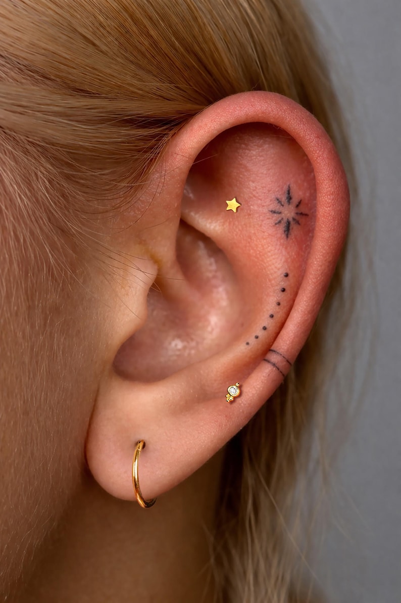 May include: An ear adorned with three gold earrings: a small star stud, a decorative stud with a clear stone, and a plain hoop. The ear features black ink tattoos including a starburst, a curved line of dots, and two parallel horizontal lines.