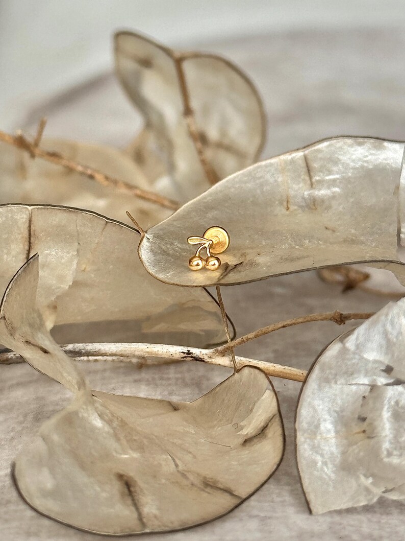 May include: A small gold stud earring shaped like two cherries with a stem. The jewelry piece features a polished metallic finish and a flat circular backing, designed as a delicate accessory for ear piercings with a simple and classic aesthetic.