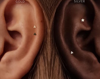 Könnte beinhalten: Zwei Ohren mit Schmuck. Das linke Ohr mit der Aufschrift GOLD zeigt zwei dreieckige Ohrstecker, einen mit klaren und einen mit schwarzen Steinen. Das rechte Ohr mit der Aufschrift SILVER zeigt einen sternförmigen und einen dreieckigen Ohrstecker.
