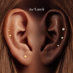 May include: Close-up of two ears, one light-skinned and one dark-skinned, each adorned with small stud earrings. The light-skinned ear has three gold studs, while the dark-skinned ear has three silver studs. The text "the Lasvit" is in the upper right corner.