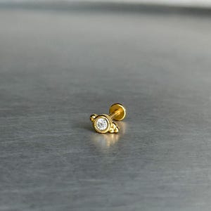 May include: A gold-toned flat back stud earring featuring a central round clear gemstone set in a bezel. The setting includes small decorative gold spheres on the sides and a thin post extending from the back, designed for body piercing jewelry.